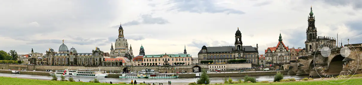 Die Elbe fließt ruhig in Dresden, während am Ufer die beeindruckende Altstadtsilhouette mit ihren markanten Kirchtürmen und historischen Gebäuden erstrahlt. Schiffe legen am Kai an und zahlreiche Menschen flanieren entlang des Flusses, was eine lebendige und geschichtsträchtige Atmosphäre schafft. Das Panorama fängt die majestätische Architektur und die idyllische Flusslandschaft dieser Kulturstadt ein.