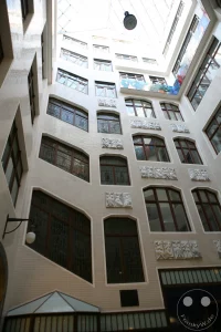 Die detailreiche Innenfassade des historischen Lichthofes A im Messepalast Specks Hof zeigt kunstvolle architektonische Elemente der Jahrhundertwende. Die hell geflieste Wandfläche ist mit markanten Kassettenfenstern, kunstvollen Bleiglasfenstern und plastischen weißen Steinreliefs verziert, die mythologische und handwerkliche Motive darstellen. Im Erdgeschoss schmiegt sich ein kleiner, traditioneller Kiosk mit verglaster Front in die Gebäudeecke, während eine klassische Wandlaterne und ein historischer Fliesenspiegel die nostalgische Atmosphäre der ältesten erhaltenen Ladenpassage Leipzigs unterstreichen.