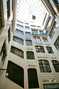 Die detailreiche Innenfassade des historischen Lichthofes A im Messepalast Specks Hof zeigt kunstvolle architektonische Elemente der Jahrhundertwende. Die hell geflieste Wandfläche ist mit markanten Kassettenfenstern, kunstvollen Bleiglasfenstern und plastischen weißen Steinreliefs verziert, die mythologische und handwerkliche Motive darstellen. Im Erdgeschoss schmiegt sich ein kleiner, traditioneller Kiosk mit verglaster Front in die Gebäudeecke, während eine klassische Wandlaterne und ein historischer Fliesenspiegel die nostalgische Atmosphäre der ältesten erhaltenen Ladenpassage Leipzigs unterstreichen.