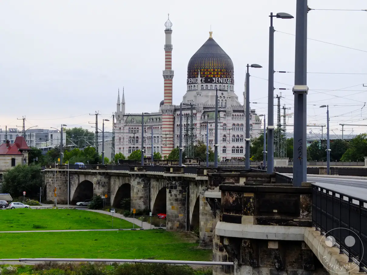 Die ehemalige Tabakfabrik Yenidze in Dresden erhebt sich imposant hinter der massiven Steinbrücke. Auf der Brücke führen Gleise und Straßen entlang, während am Ufer Grünflächen und Bäume die Szenerie auflockern. Der leicht bewölkte Himmel verleiht der Ansicht eine ruhige und alltägliche Atmosphäre.
