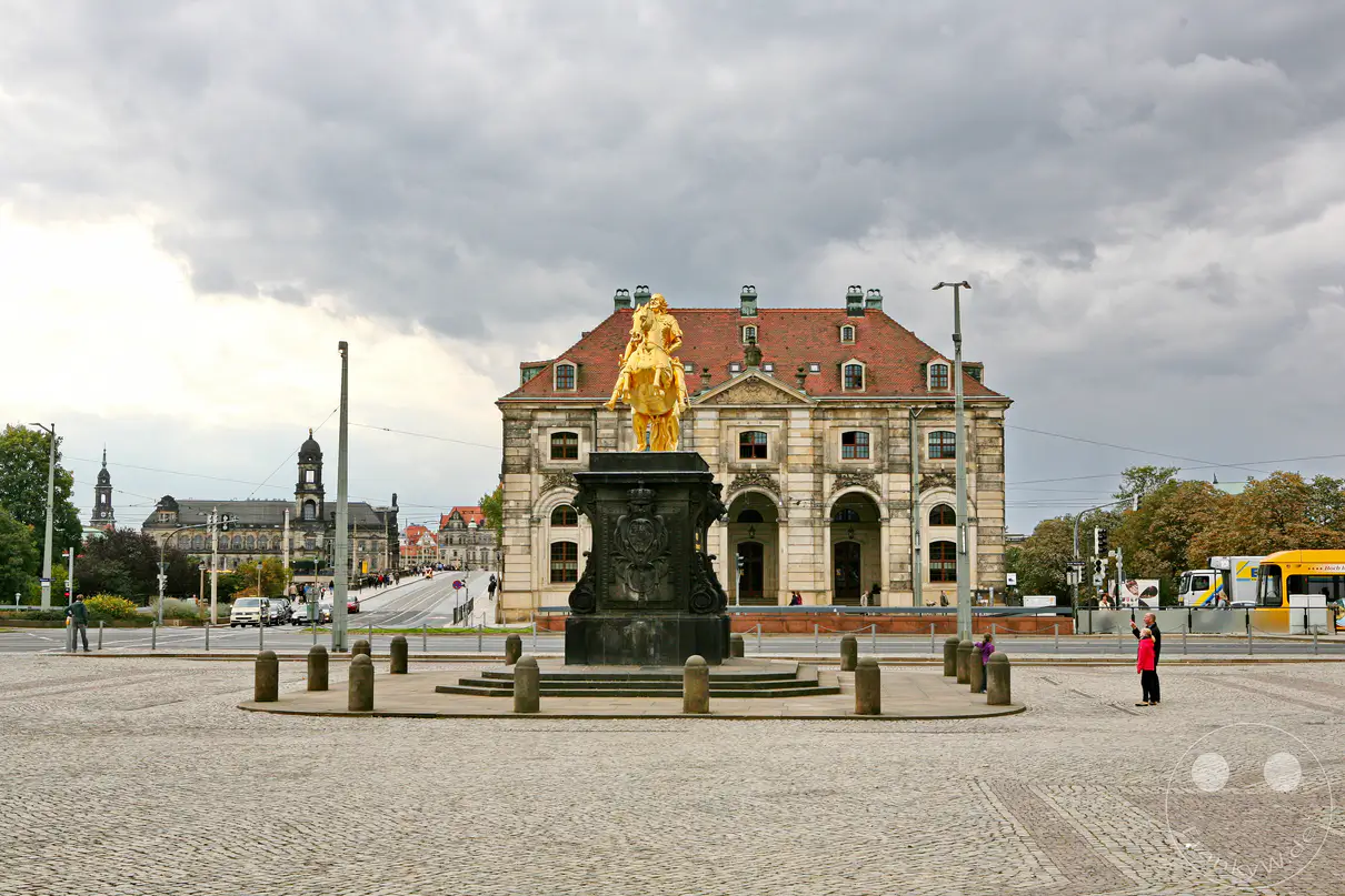 Das Reiterstandbild von Augustus dem Starken thront majestätisch auf dem Neumarkt in Dresden, Deutschland. Die prachtvolle barocke Architektur, die teilweise im Hintergrund zu sehen ist, bildet eine beeindruckende Kulisse für das goldene Reiterstandbild. Die Szene wird von einem dramatisch bewölkten Himmel überlagert, der der Aufnahme eine melancholische und gleichzeitig monumentale Atmosphäre verleiht.