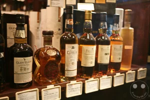 Eine edle Auswahl hochwertiger schottischer Single Malt Whiskys präsentiert sich ordentlich aufgereiht in einem hölzernen Verkaufsregal in der Ladenpassage Specks Hof. Im Vordergrund stehen charakteristische Flaschen bekannter Destillerien wie Glen Elgin 12 Jahre, Cardhu sowie eine Abfüllung von Clynelish, gefolgt von mehreren markanten Editionen der Brennerei Talisker. Kleine, weiße Informations- und Preisschilder an der Regalkante weisen die spezifischen Details für Whiskyliebhaber aus und unterstreichen das exklusive Genusssortiment des Fachgeschäfts.