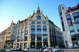 Die prachtvolle, reich mit Goldschmuck verzierte Sandsteinfassade des historischen Kaufhauses Ebert am Leipziger Thomaskirchhof präsentiert sich im hellen Sonnenlicht. Das zwischen 1903 und 1904 im prägnanten Jugendstil errichtete Eckgebäude besticht durch seine geschwungenen Fensterbögen, monumentalen Giebelstrukturen und den kunstvoll verzierten Turmaufbau an der Dachspitze. Im Erdgeschoss des denkmalgeschützten Bauwerks befindet sich heute eine Filiale der Commerzbank, während im Vordergrund parkende Autos, Fahrräder und Passanten das urbane Leben auf dem gepflasterten Platz widerspiegeln.
