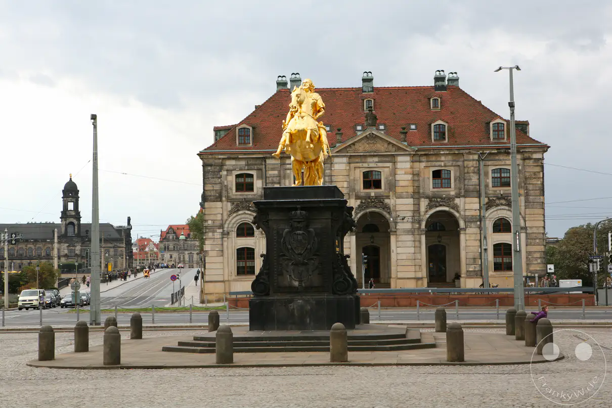 Das weltberühmte Reiterstandbild des Goldenen Reiters steht als glänzendes Denkmal im Zentrum des Neustädter Marktes in Dresden. Das Monument zeigt den sächsischen Kurfürsten und polnischen König August den Starken in römischer Rüstung auf einem steigenden Lipizzanerhengst in Richtung Polen reiten. Direkt dahinter erhebt sich das historische Neustädter Blockhaus, das im 18. Jahrhundert nach Entwürfen von Zacharias Longuelune als Wachgebäude errichtet wurde. Auf der linken Seite öffnet sich der Blick zur Augustusbrücke und der Altstadtsilhouette, während vereinzelte Passanten den weitläufigen, gepflasterten Platz unter einem wolkenverhangenen Himmel überqueren.