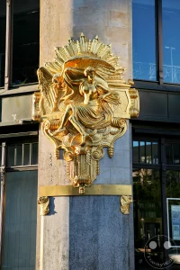 Ein kunstvoll gearbeitetes, goldglänzendes Relief ziert als markanter Bauschmuck eine Sandsteinsäule an der Außenfassade des historischen Kaufhauses Ebert und zeigt die Allegorie der Genusssucht (Luxuria). Die dynamisch gestaltete Jugendstil-Plastik stellt eine schwebende weibliche Gestalt dar, die von schweren, elegant geschwungenen Stofffalten umgeben ist. Das im warmen Sonnenlicht leuchtende Kunstwerk bildete bei der Eröffnung des luxuriösen Damenbekleidungshauses im Jahr 1904 zusammen mit seinem Gegenüber ein augenzwinkerndes Symbol für den weltlichen Konsum und bildet ein prachtvolles historisches Detail am Eingang des heutigen Bankgebäudes.