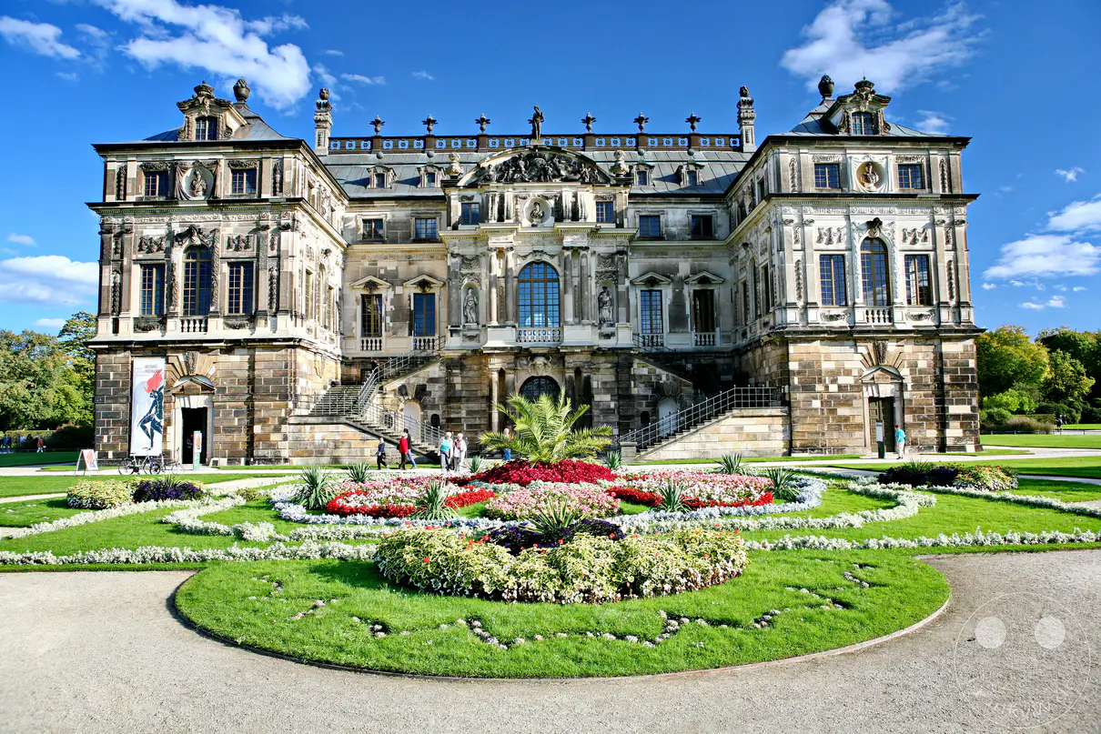 Das Palais im Großen Garten in Dresden, ein prächtiges Lustschloss im barocken Stil, erhebt sich majestätisch vor einem blauen Himmel mit weißen Wolken. Ein bunter, kunstvoll angelegter Blumen- und Gräsergarten bildet den Vordergrund und lädt zu einem Spaziergang durch die gepflegten Anlagen ein. Die Szenerie strahlt eine heitere und königliche Atmosphäre aus, die den Charme des historischen Ortes unterstreicht.