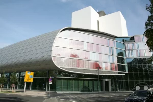Der Erweiterungsbau der Deutschen Nationalbibliothek in Leipzig präsentiert sich mit einer auffälligen, geschwungenen Glasfassade, die von roten und weißen Elementen akzentuiert wird. Bäume und der blaue Himmel spiegeln sich in den großen Glasflächen wider und erzeugen eine helle und luftige Atmosphäre.