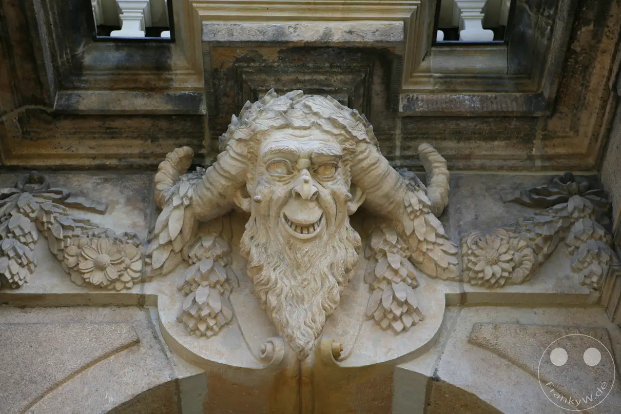 Ein grinsender Pan-Kopf mit markanten Hörnern und langem Bart prangt als steinerner Neidkopf oder Mascheron über einem Portalbogen des frühbarocken Palais im Großen Garten. Die detailreiche Sandsteinskulptur ist kunstvoll in eine Girlande aus Blüten und Blättern eingebunden, die sich zu beiden Seiten entlang des Sims erstreckt. Oberhalb der fratzenhaften Maske sind die Ansätze von Fensteröffnungen und weißen Balustraden des historischen Lustschlosses zu erkennen. Solche mythologischen Fratzen und heroischen Reliefs zieren die gesamte Außenfassade des Prachtbaus und spiegeln den typischen Dekorationsstil des sächsischen Barocks wider.
