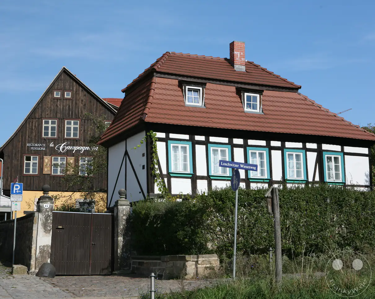 Das historische, denkmalgeschützte Fährhäuschen im Dresdner Stadtteil Loschwitz präsentiert sich an einem sonnigen Tag mit einer charakteristischen weiß-schwarzen Fachwerkfassade, türkisfarbenen Fensterrahmen und einem roten Ziegeldach. Direkt dahinter ragt das dunkle, holzverkleidete Gebäude des traditionsreichen Ristorante "La Campagnola" empor, während ein blaues Straßenschild den "Loschwitzer Wiesenweg" ausweist. Die idyllische Szenerie an der Elbe vermittelt eine einladende, dörfliche Ruhe inmitten der sächsischen Landeshauptstadt in Deutschland.