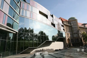 Der Erweiterungsbau der Deutschen Nationalbibliothek in Leipzig präsentiert sich mit einer auffälligen, geschwungenen Glasfassade, die von roten und weißen Elementen akzentuiert wird. Bäume und der blaue Himmel spiegeln sich in den großen Glasflächen wider und erzeugen eine helle und luftige Atmosphäre.