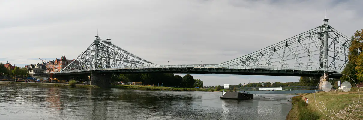 Die Loschwitzer Brücke, auch bekannt als "Blaues Wunder", überspannt majestätisch die Elbe in Dresden, Deutschland. Die markante Stahlkonstruktion bildet zusammen mit den umliegenden Gebäuden und der ruhigen Flusslandschaft ein beeindruckendes Stadtpanorama an einem bewölkten Tag. Schiffe passieren die Brücke, während auf dem Uferweg Menschen flanieren und Bauarbeiten auf der linken Seite stattfinden.