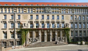 Das prächtige Gebäude der Deutschen Nationalbibliothek in Leipzig präsentiert sich mit einer beeindruckenden neobarocken Architektur unter einem roten Ziegeldach. Die Fassade ist reich verziert mit Skulpturen, Friesen und einem großen Zifferblatt einer Uhr, was die historische Bedeutung unterstreicht. Die Szene strahlt eine Atmosphäre von Bildung und kulturellem Erbe aus, verstärkt durch das klare blaue Himmelslicht, das die Details der aufwendigen Bauweise betont.