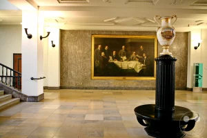 Eine historische Malerei mit mehreren Männern, die an einem Tisch sitzen, dominiert die Wand einer Eingangshalle in der Deutschen Nationalbibliothek in Leipzig, Deutschland. Eine beeindruckende Vase auf einem Sockel steht prominent im Vordergrund und spiegelt die architektonische Umgebung wider. Die Szene vermittelt eine Atmosphäre von Bedeutung und historischem Erbe, verstärkt durch die klassische Architektur und die gedämpfte Beleuchtung.