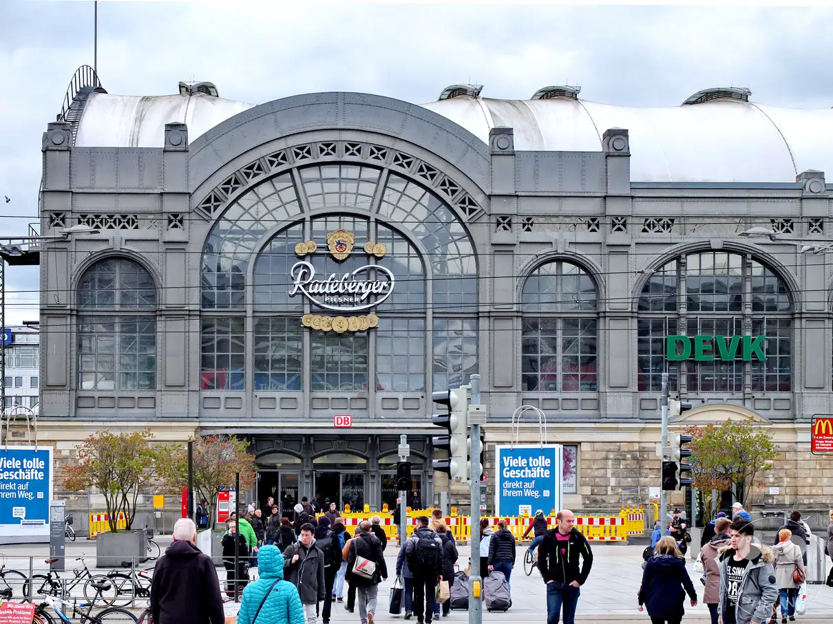 Die imposante Fassade des Dresdner Hauptbahnhofs mit dem Schriftzug "Radeberger Pilsner" und einem "DB"-Logo dominiert die Szene. Eine belebte Straße mit vielen Menschen, Fahrrädern und einer Straßenbahnhaltestelle belebt den Vordergrund, während im Hintergrund Geschäfte zu erkennen sind. Die Architektur des Bahnhofs und das geschäftige Treiben der Stadt erzeugen eine dynamische Atmosphäre.