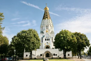 Die Russische Gedächtniskirche des Heiligen Alexej in Leipzig, Deutschland, präsentiert sich an einem sonnigen Tag mit ihrem markanten goldenen Turm und weißer Fassade. Umrahmt von üppigen grünen Bäumen strahlt die orthodoxe Kirche eine feierliche und ruhige Atmosphäre aus. Im Hintergrund sind Bäume und eine Straße mit parkenden Autos zu sehen, die das Bild einer belebten, aber friedlichen Umgebung vermitteln.