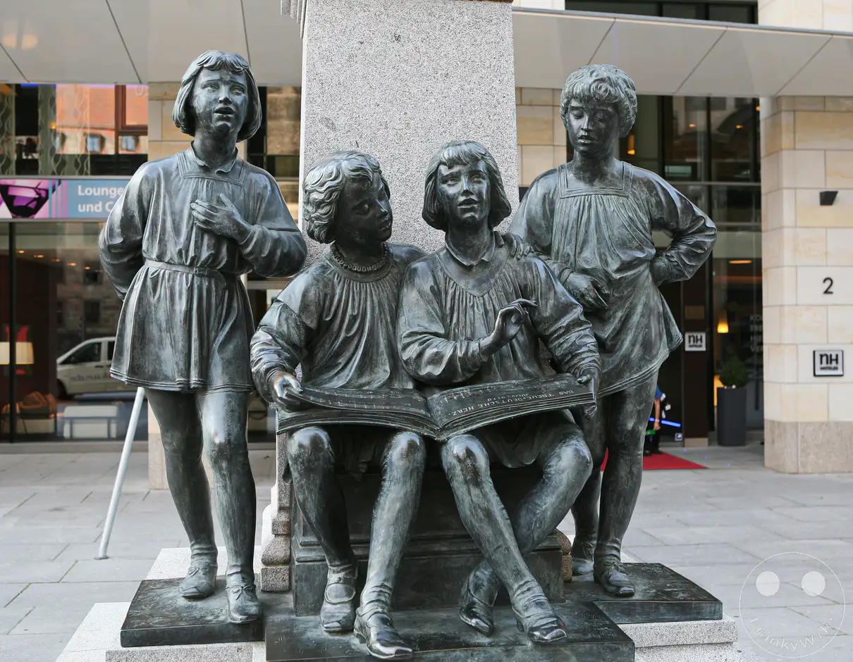 Eine bronzene Skulptur in Dresden, Deutschland, zeigt vier Kinderfiguren, die auf einem Podest sitzen und stehen. Zwei der sitzenden Kinder lesen gemeinsam aus einem aufgeschlagenen Buch, während die anderen beiden stehenden Kinder sie beobachten. Die detailreiche Darstellung vermittelt eine Atmosphäre der Gemeinschaft und des Lernens.