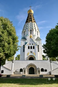 Die Russische Gedächtniskirche des Heiligen Alexej in Leipzig, Deutschland, präsentiert sich an einem sonnigen Tag mit ihrem markanten goldenen Turm und weißer Fassade. Umrahmt von üppigen grünen Bäumen strahlt die orthodoxe Kirche eine feierliche und ruhige Atmosphäre aus. Im Hintergrund sind Bäume und eine Straße mit parkenden Autos zu sehen, die das Bild einer belebten, aber friedlichen Umgebung vermitteln.