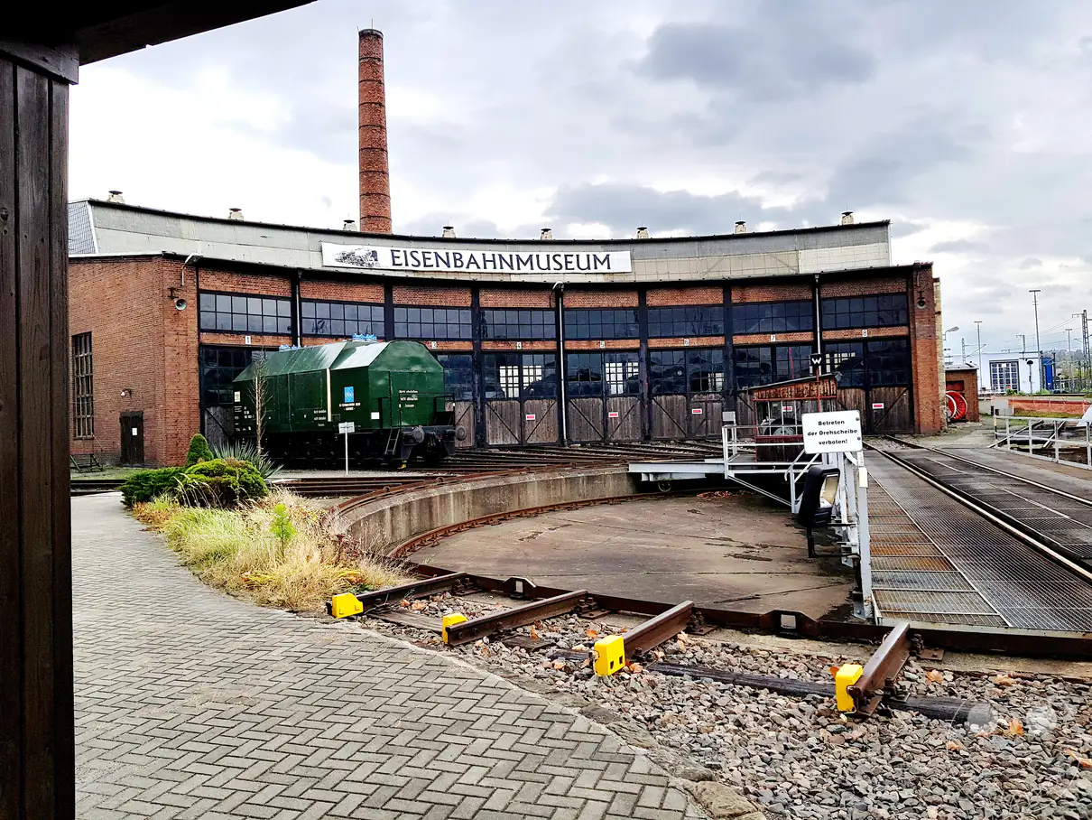 Das Eisenbahnmuseum in Dresden präsentiert sich an einem bewölkten Tag mit seiner charakteristischen runden Lokschuppenanlage und dem hohen Schornstein. Ein grüner Güterwagen steht auf dem Drehschemel, umgeben von einem komplexen Schienennetz und gepflasterten Wegen. Die Szenerie strahlt eine Atmosphäre der Industriegeschichte und Nostalgie aus, eingebettet in eine graue, herbstliche Wetterlage.