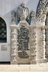 Die detailreiche steinerne Gedenktafel an der Außenwand der Russischen Gedächtniskirche erinnert an die Gefallenen der historischen Völkerschlacht bei Leipzig im Jahr 1813. Das monumentale Relief wird von einem großen russischen Doppeladler gekrönt und ist mit einer ausführlichen deutschsprachigen Inschrift versehen, welche die Truppenstärken sowie die genauen Verlustzahlen der verbündeten Nationen dokumentiert. Flankiert von zwei verzierten, pyramidenförmigen Steinsäulen schließt das historische Denkmal direkt an das reich ornamentierte Portal der Kirche des Heiligen Alexij an, die als Ehrenmal für die gefallenen russischen Soldaten errichtet wurde.