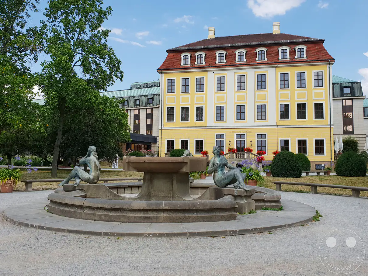 Zwei bronzene Figurenskulpturen ruhen am Rand eines runden Steingartens im Außenbereich vor einem prachtvollen, barocken Barockpalais in Dresden. Das historische, gelb gestrichene Gebäude mit seinen symmetrischen Fensterreihen und dem Mansarddach bildet die Kulisse für diese elegante Parkanlage in Deutschland. Eine ruhige, herrschaftliche und historisch anmutende Stimmung prägt die Szene im Garten des heutigen Bilderberg Bellevue Hotels.