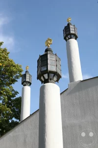 Drei aufwendig gestaltete Laternen auf weißen Säulen ragen vor einem strahlend blauen Himmel empor. Die Architektur der Russischen Gedächtniskirche des Heiligen in Leipzig präsentiert sich mit markanter Textur an ihrer Fassade. Ein Teil eines Baumes mit grünem Laubwerk ist im linken Bildbereich zu sehen und kontrastiert die kühlen Weiß- und Blautöne.