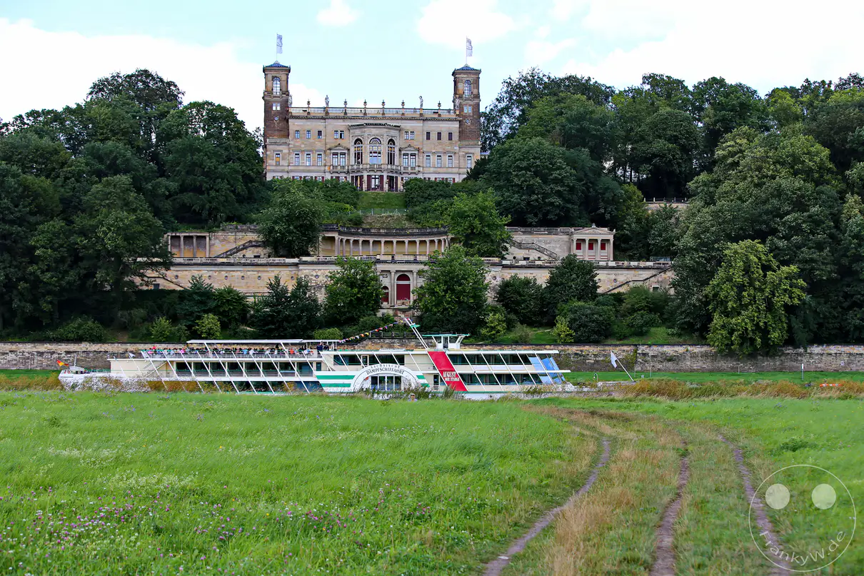 Das Schloss Albrechtsberg thront majestätisch über dem Elbufer in Dresden, Deutschland. Vor der Kulisse des Schlosses gleitet ein historisches Dampfschiff auf der Elbe, umgeben von saftig grünen Wiesen. Eine friedliche und malerische Szenerie, die die Schönheit der Landschaft und die historische Architektur vereint.
