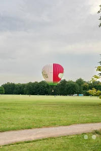 Ein bunter Heißluftballon schwebt am Himmel über der weitläufigen Parkwiese des Landschaftsparks Rosental in Leipzig. Im Hintergrund sind Bäume des Leipziger Auwalds sowie städtische Gebäude zu sehen, während Menschen entspannt die grüne Fläche genießen. Die Szenerie vermittelt eine friedliche Atmosphäre von Freizeit und Natur mitten in der Stadt.