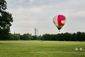 Ein bunter Heißluftballon schwebt am Himmel über der weitläufigen Parkwiese des Landschaftsparks Rosental in Leipzig. Im Hintergrund sind Bäume des Leipziger Auwalds sowie städtische Gebäude zu sehen, während Menschen entspannt die grüne Fläche genießen. Die Szenerie vermittelt eine friedliche Atmosphäre von Freizeit und Natur mitten in der Stadt.