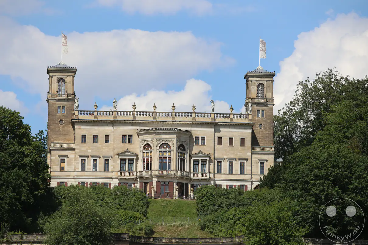 Die symmetrische Sandsteinfassade von Schloss Albrechtsberg in Dresden zeigt sich in einer Frontalansicht unter einem blau-weißen Sommerhimmel. Zwei quadratische Ecktürme mit Aussichtsplattformen und wehenden Flaggen rahmen den zentralen, mehrstöckigen Gebäudekomplex ein. Ein prachtvoller, halbrunder Mittelrisalit mit hohen Rundbogenfenstern und kunstvollen Reliefs betont das Zentrum der Schlossarchitektur. Dichte, grüne Baumkronen und üppige Hecken am unteren Bildrand umschließen den Sockelbereich des historischen Bauwerks.