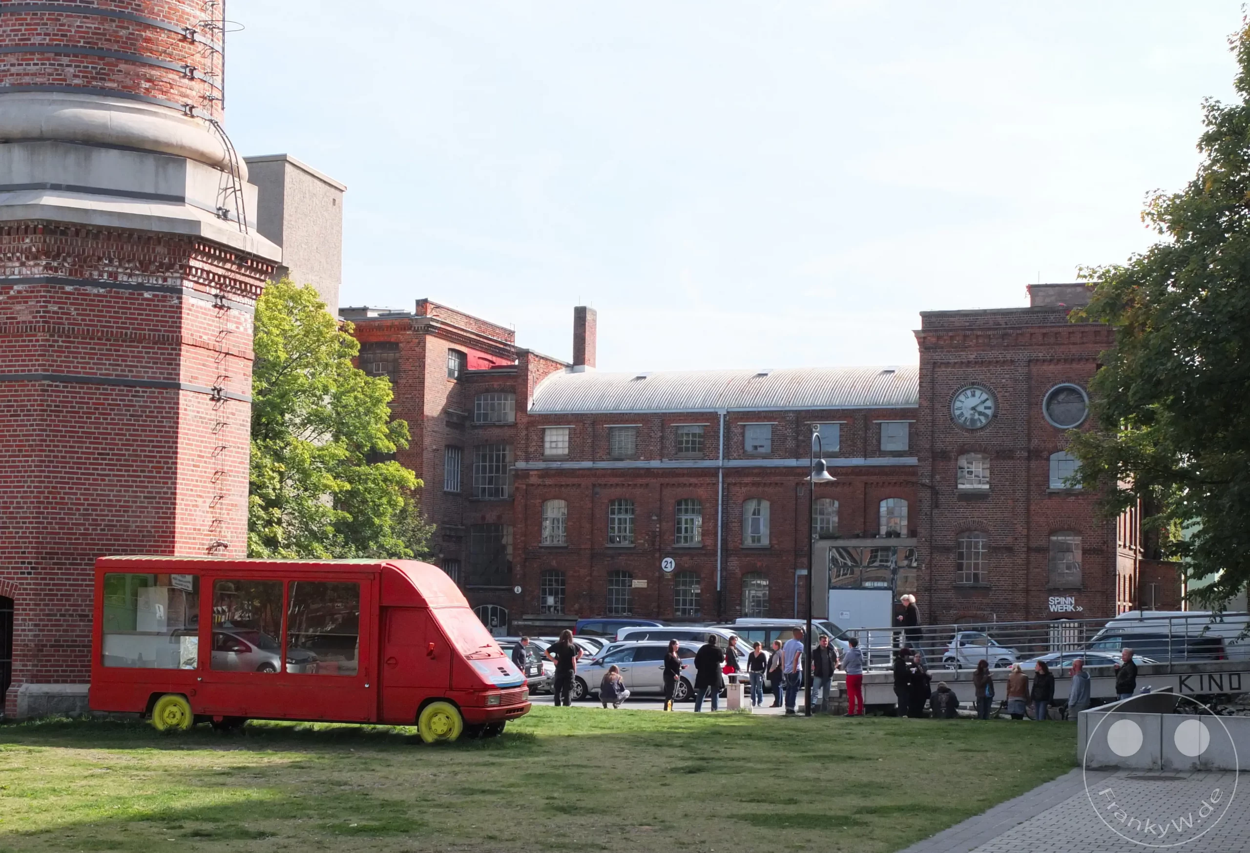 In Leipzig-Lindenau, auf dem Gelände der ehemaligen Leipziger Baumwollspinnerei, versammeln sich Menschen vor historischen Industriebauten. Ein roter, umgebauter Kleinbus steht auf einer Wiese vor dem imposanten Fabrikschornstein, während im Hintergrund weitere Gebäude und geparkte Autos zu sehen sind. Die Szene vermittelt eine Atmosphäre des urbanen Lebens und der kulturellen Nutzung historischer Industriearchitektur.