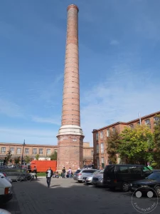 Der monumentale Fabrikschornstein der ehemaligen Leipziger Baumwollspinnerei dominiert den blauen Himmel über Leipzig Lindenau. Industriebauten im Hintergrund und parkende Autos am Straßenrand zeugen vom urbanen Charakter des Viertels, das heute ein lebendiges Kunst- und Kulturzentrum ist. Ein Mann spaziert auf dem Gehweg, während Menschen vor einem Gebäude verweilen und die sonnige Atmosphäre genießen.