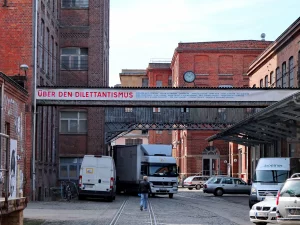 In Leipzig-Lindenau, auf dem Gelände der ehemaligen Leipziger Baumwollspinnerei, präsentiert ein Banner die Namen von Künstlern und die Ausstellung „Über den Dilettantismus“. Die Szenerie, geprägt von industrieller Backsteinarchitektur und gepflasterten Wegen, vermittelt eine Atmosphäre von Kreativität und urbanem Leben, ergänzt durch Fahrzeuge und Fahrräder, die auf Aktivitäten in diesem Kunst- und Kulturzentrum hindeuten.