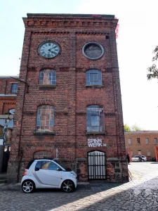 In Leipzig, im Stadtteil Lindenau, präsentiert sich die ehemalige Leipziger Baumwollspinnerei als lebendiges Kunst- und Kulturzentrum. Der markante Uhrturm des Spinnwerks dominiert die Ansicht, während ein weißes Smart-Fahrzeug davor parkt und ein Teil des historischen Kopfsteinpflasters sichtbar ist. Die Szene strahlt eine industrielle Vergangenheit aus, die durch moderne Nutzung als kreativer Ort belebt wird.