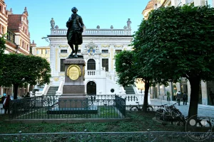 Das Goethedenkmal auf dem Naschmarkt in Leipzig steht prominent vor der beeindruckenden Fassade des Alten Rathauses am Augustusplatz. Die Szene wird durch die späte Nachmittagssonne beleuchtet, die eine ruhige und historische Atmosphäre schafft. Neben dem Denkmal und dem historischen Gebäude sind gepflegte Grünflächen und einige Bäume zu sehen, die das städtische Bild ergänzen.