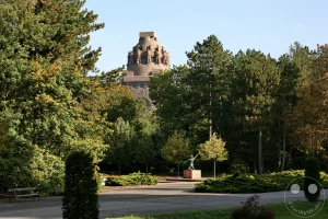 In Leipzig, Deutschland, ragt das imposante Völkerschlachtdenkmal hinter einem Park hervor, der mit Bäumen und Sträuchern geschmückt ist. Im Vordergrund steht eine Bronzestatue auf einem Sockel, die eine mahnende Geste in Richtung des Denkmals ausführt. Die friedliche Parklandschaft mit dem ernsten Monument schafft eine nachdenkliche Atmosphäre an einem klaren Tag.