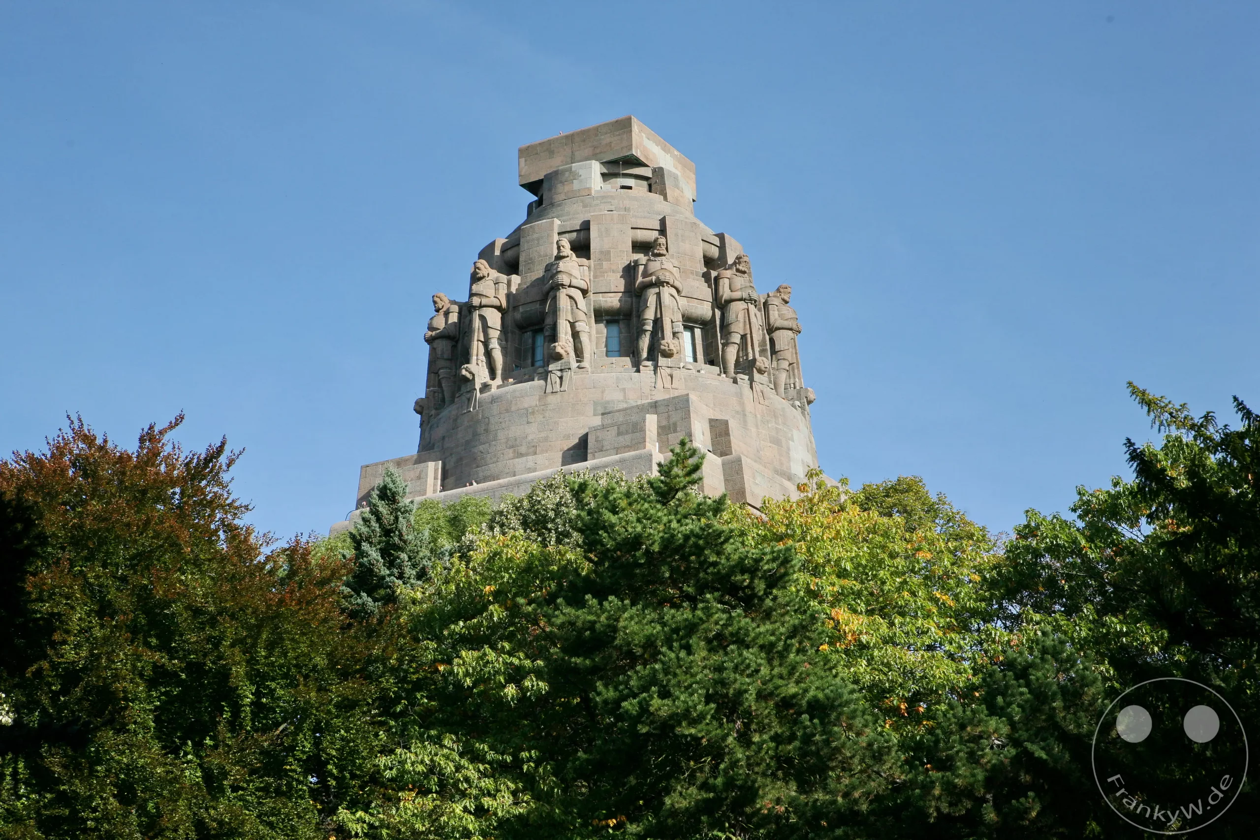 Das Völkerschlachtdenkmal in Leipzig, Deutschland, ragt majestätisch in den blauen Himmel. Bewundernswerte Skulpturen von Kriegern krönen den steinernen Koloss, umgeben von üppigem grünen Laub. Die monumentale Architektur und die historischen Statuen strahlen eine erhabene und geschichtsträchtige Atmosphäre aus.