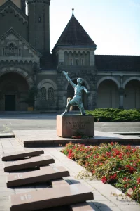 Die eindrucksvolle Bronzeplastik des Aufsteigenden von Walter Arnold erhebt sich im Zentrum des Ehrenhains für die Opfer des Nationalsozialismus auf dem Leipziger Südfriedhof. Die ausdrucksstarke Skulptur eines Mannes mit erhobenem Arm steht auf einem steinernen Sockel, der die eingravierte Inschrift „Zum Tode geführt und siehe wir leben“ trägt. Im Vordergrund säumen flache Gedenktafeln aus rötlichem Granit und ein herbstlich blühendes Rosenbeet den gepflasterten Vorplatz, während im Hintergrund die monumentale neuromanische Kapellenanlage des Friedhofs aufragt.