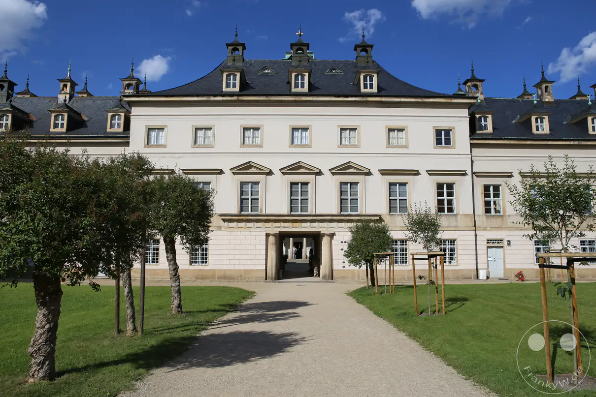 Die symmetrische Fassade des Bergpalais von Schloss Pillnitz in Dresden zeigt sich unter einem blauen Himmel mit vereinzelten weißen Wolken. Ein breiter, heller Kiesweg führt im Vordergrund direkt auf das zentrale Säulenportal des historischen Schlossgebäudes zu. Das helle Bauwerk besticht durch ein ausladendes, dunkles Mansarddach, welches mit kunstvollen Gauben und kleinen Turmspitzen verziert ist. Gepflegte grüne Rasenflächen und mehrere junge, gestützte Bäume flankieren den Zugangsweg auf beiden Seiten.