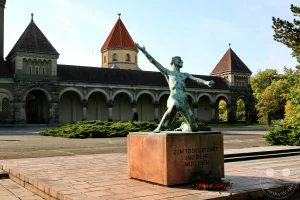 Die eindrucksvolle Bronzeplastik des Aufsteigenden von Walter Arnold erhebt sich im Zentrum des Ehrenhains für die Opfer des Nationalsozialismus auf dem Leipziger Südfriedhof. Die ausdrucksstarke Skulptur eines Mannes mit erhobenem Arm steht auf einem steinernen Sockel, der die eingravierte Inschrift „Zum Tode geführt und siehe wir leben“ trägt. Im Vordergrund säumen flache Gedenktafeln aus rötlichem Granit und ein herbstlich blühendes Rosenbeet den gepflasterten Vorplatz, während im Hintergrund die monumentale neuromanische Kapellenanlage des Friedhofs aufragt.