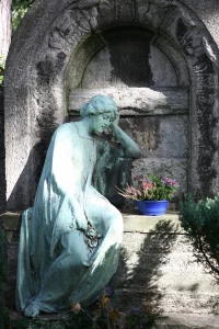 Eine gartendenkmalpflegerisch wertvolle, patinierte Bronzeskulptur einer trauernden Frau lehnt sich schwermütig an eine massive steinerne Grabstele auf dem Leipziger Südfriedhof. Die lebensgroße, im klassischen Jugendstil gestaltete Frauenfigur sitzt in fließendem Gewand barfuß am Sockel der historischen Familiengrabstätte und stützt ihren gesenkten Kopf nachdenklich in die linke Hand. Eine blaue Pflanzschale mit herbstlich blühender Heide und bunten Blumen setzt einen lebendigen Akzent vor der verwitterten Architektur des geschichtsträchtigen Grabmals, das von schattenspendenden Nadelgehölzen umrahmt wird.