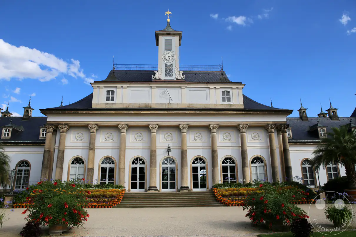 Die symmetrische, hell verputzte Fassade des Neuen Palais von Schloss Pillnitz in Dresden präsentiert sich im Zentrum des Fliederhofs unter einem blauen Himmel. Hohe Säulen im klassizistischen Stil tragen das ausladende, dunkle Mansarddach, aus dessen Mitte ein schlanker weißer Uhrenturm emporragt. Eine große, filigran gezeichnete Sonnenuhr schmückt die Wandfläche oberhalb der zentralen Fensterreihen und unterhalb des Turmansatzes. Mehrere Treppenstufen führen im Vordergrund zu den großen Rundbogenportalen hinauf, flankiert von bunt bepflanzten Blumenbeeten und dekorativen Pflanzkübeln auf dem weiten Kiesplatz.