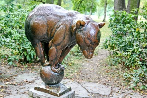 Eine detaillierte Bronzeplastik eines Bären, der auf einer Kugel balanciert, steht inmitten eines herbstlichen Gartens im Clara-Zetkin-Park in Leipzig, Deutschland. Die Skulptur aus wettergegerbtem Metall fängt die Kraft und Anmut des Tieres ein, umgeben von üppigem Grün und gefallenem Laub. Die Szene vermittelt eine ruhige und nachdenkliche Atmosphäre, die die Kunstwerke in ihrer natürlichen Umgebung hervorhebt.