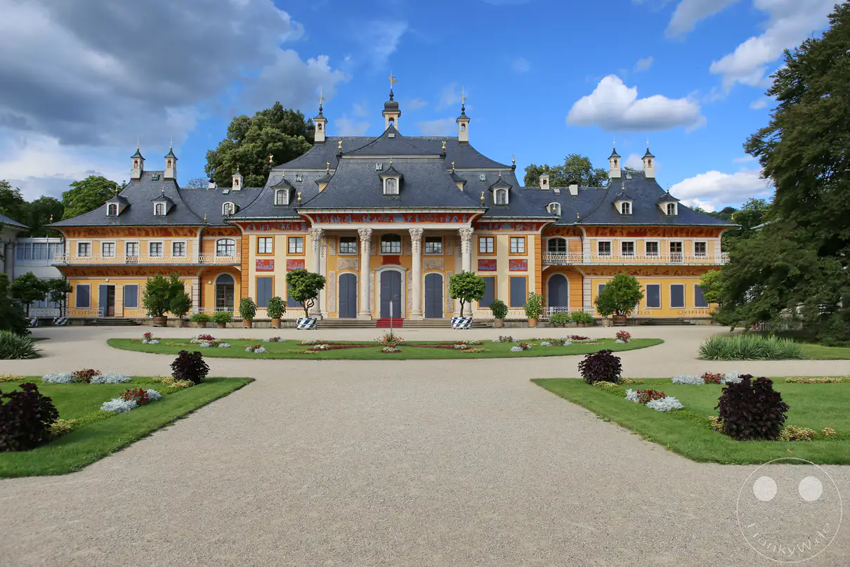 Die prächtige, ockerfarbene Fassade des Wasserpalais von Schloss Pillnitz in Dresden erstreckt sich hinter einem symmetrisch angelegten Barockgarten unter einem blau-weißen Wolkenhimmel. Ein zentraler Portikus mit hohen Säulen und chinesisch anmutenden Dachschwüngen prägt die Architektur des historischen Lustschlosses. Ein breiter, heller Kiesweg führt im Vordergrund direkt auf die kunstvoll gestalteten Blumenbeete und Rasenflächen vor dem Gebäude zu. Dichte, dunkelgrüne Baumkronen rahmen das gesamte barocke Schlossareal auf der rechten Seite harmonisch ein.
