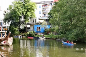 Ein sonniger Tag an der Weißen Elster in Leipzig, Deutschland, wo Menschen in einem blauen Boot die ruhige Atmosphäre des Kanals genießen. Am Ufer liegen kleinere Boote und bunte Häuser, umgeben von üppigem Grün und Bäumen, was eine friedliche Stadtszenerie schafft. Die Reflektionen im Wasser spiegeln die lebhafte Kulisse und die entspannte Stimmung des Ortes wider.