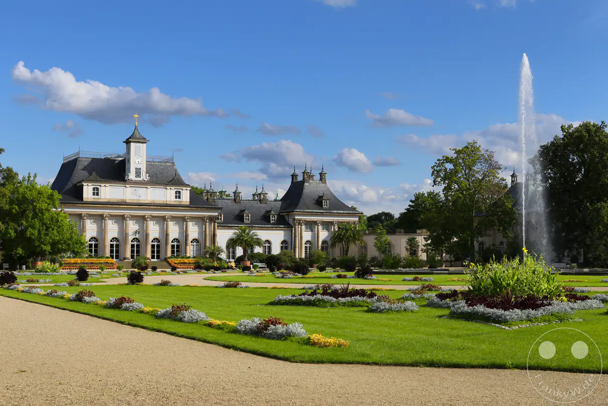 Die hell verputzten Gebäudeflügel des Neuen Palais von Schloss Pillnitz in Dresden erstrecken sich im Hintergrund unter einem blau-weißen Sommerhimmel. Vor dem historischen Schlosskomplex breitet sich der weitläufige Lustgarten mit geometrisch angeordneten Rasenflächen und bunt blühenden Blumenrabatten aus. Auf der rechten Seite schießt eine hohe, vertikale Wasserfontäne aus einem großen Brunnenbecken empor und bildet einen dynamischen Blickfang. Ein geschwungener, heller Kiesweg im linken Vordergrund führt Spaziergänger durch die weitläufige, barocke Parkanlage.