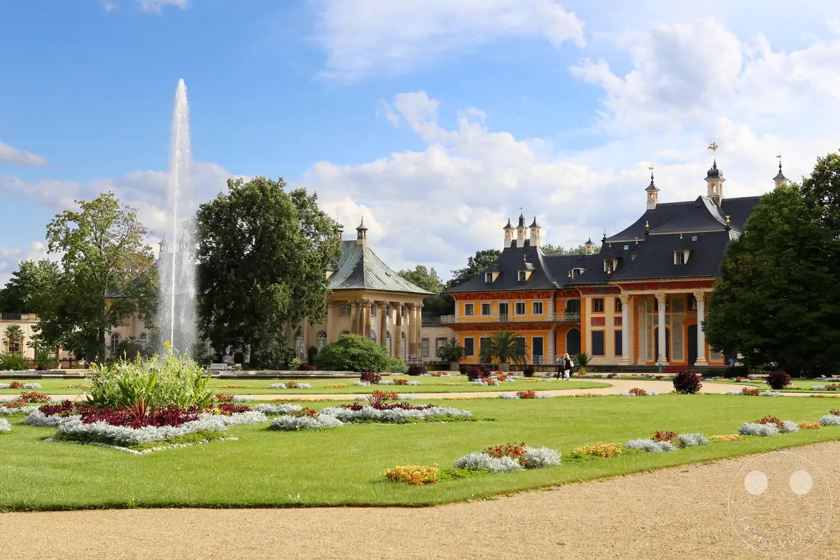 Die prächtige Schlossanlage von Schloss Pillnitz in Dresden erstreckt sich auf der rechten Seite hinter einer weitläufigen Parkanlage unter einem blau-weißen Sommerhimmel. Die ockerfarbenen Fassaden des Wasserpalais und der angrenzenden Pavillons zeichnen sich durch geschwungene, dunkle Dächer im chinesischen Stil aus. Auf der linken Seite schießt eine mächtige vertikale Wasserfontäne aus einem großen, reich bepflanzten Brunnenbeet empor. Im Vordergrund führen geschwungene Kieswege entlang geometrisch angeordneter Rasenflächen mit dekorativen, bunt blühenden Blumenrabatten.