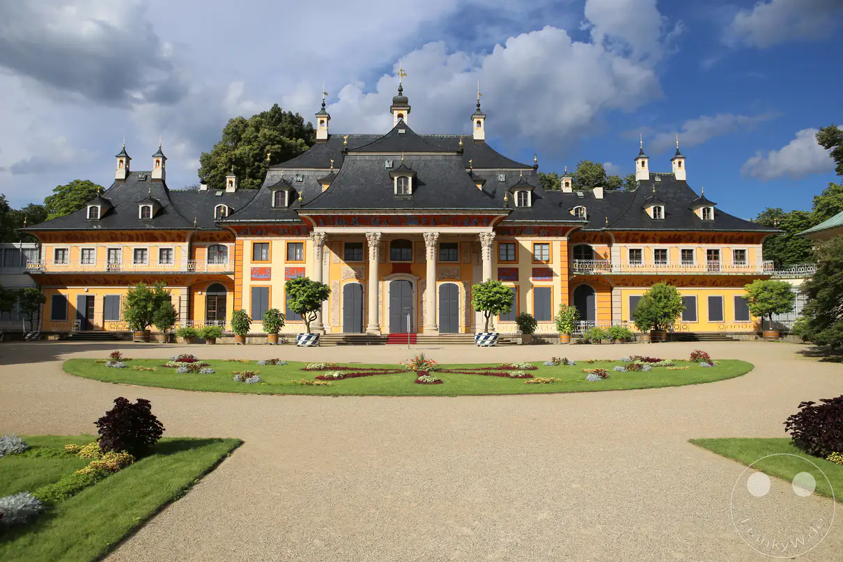 Die prächtige, ockerfarbene Frontfassade des Bergpalais von Schloss Pillnitz in Dresden erstreckt sich hinter einer symmetrisch angelegten Gartenanlage unter einem blau-weißen Wolkenhimmel. Ein zentraler Portikus mit hohen Säulen und chinesisch geschwungenen Dachelementen prägt die Architektur des historischen Lustschlosses. Ein breiter, heller Kiesweg führt im Vordergrund direkt auf die kunstvoll gestalteten Rasenflächen und Blumenbeete vor dem Gebäude zu. Mehrere kleine, dekorative Bäume in Pflanzkübeln sowie dichte grüne Baumkronen im Hintergrund rahmen das barocke Schlossareal harmonisch ein.