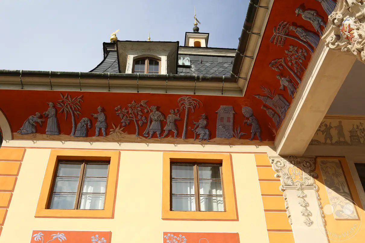 Die ockerfarbene Außenwand eines Gebäudeflügels von Schloss Pillnitz in Dresden zeigt aufwendige barocke Dekorationen unter einem hellen Himmel. Ein breiter, rötlicher Fries zieht sich direkt unterhalb der Dachrinne entlang und ist mit einer Vielzahl filigraner, blau-grauer Figurenreliefs im chinesischen Stil verziert. Zwei quadratische Holzfenster mit warmtonigen Rahmen strukturieren die darunter liegende, glatt verputzte Wandfläche auf der linken Seite. Ein dunkles Mansarddach mit einer verzierten Dachgaube und einer kleinen Turmspitze ragt im oberen Bilddrittel hinter der Fassadenkante empor.