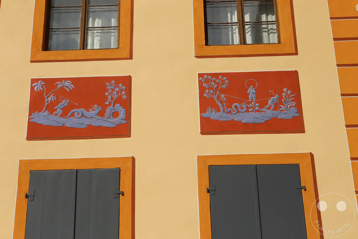 Ein Ausschnitt der ockerfarbenen Fassade an einem der Palais von Schloss Pillnitz in Dresden zeigt kunstvolle architektonische Details. Zwischen den oberen Sprossenfenstern und den unteren, geschlossenen grauen Fensterläden befinden sich zwei rechteckige, rötlich grundierte Wandflächen. Diese Felder sind mit filigranen, blau-grauen Malereien im Stil der Chinoiserie verziert, die asiatisch anmutende Figuren und exotische Pflanzenmotive darstellen. Die Fensterrahmen und die glatten Wandflächen werden durch farblich abgesetzte, orangefarbene Umrandungen dekorativ hervorgehoben.