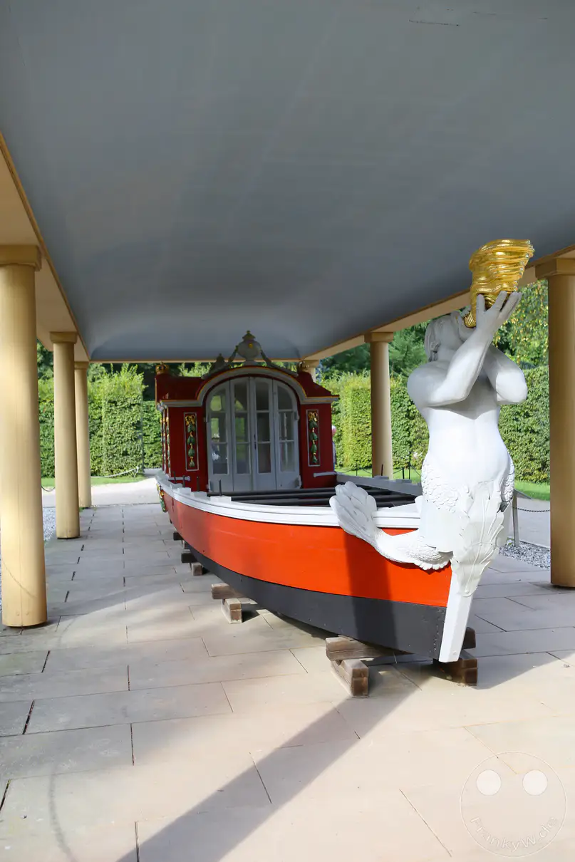 Die historische Tritonengondel steht auf Holzbalken aufgebockt unter einem schützenden, säulengestützten Pavillondach im Schlosspark von Pillnitz. Das prunkvolle barocke Boot besitzt einen rot-schwarz gestrichenen Rumpf und ein verziertes, geschlossenes Kabinenhäuschen im hinteren Bereich. Am Bug der Gondel ragt eine auffällige, weiße Skulptur eines Tritons empor, der aus einem goldenen Füllhorn bläst. Im Hintergrund erstrecken sich die gepflegten, hohen Hecken und Grünanlagen des königlichen Schlossgartens entlang des gepflasterten Bodens.